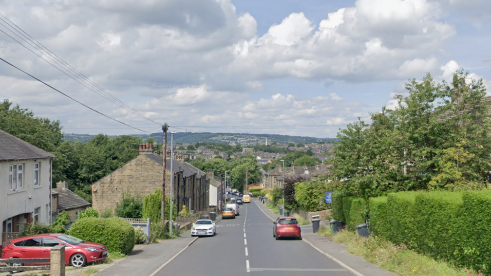 A residential street overlooking hills with cars parked alongside hedges, trees and houses.