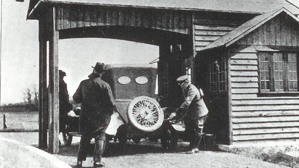 A black and white image shows a uniformed AA patrolman wearing a cap filling a car with fuel. The car sits under a wooden canopy next to a wooden single-storey building with a window. To the side of the car a man stands with his back to the camera. He is wearing a long dark coat over dark trouser and a dark hat with a brim. A third man wearing a flat-cap style hat can be seen to the rear of the wooden canopy, on the left side behind the front end of the car.