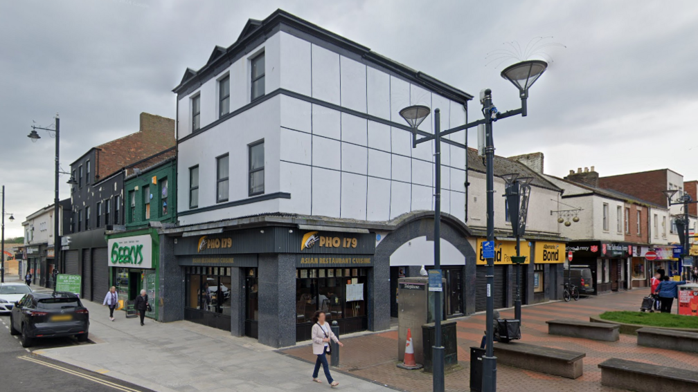 Photo of two-storey building above a restaurant which says "Pho 179". Next to the restaurant is a place called "Geekys". The two-storey building is white and has three windows on each floor. The walls are white. There is street lighting in front of it.