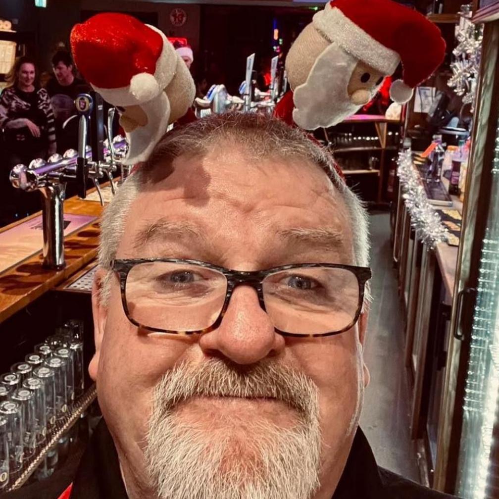 A man with a white beard wearing glasses, he has a Christmas head band on with miniature Santa's. The picture is a selfie he's taken behind the bar of the pub.