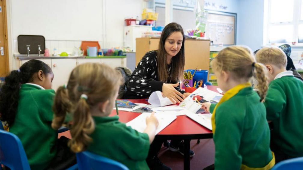 A primary school teacher sitting with a group of young children in green school jumpers at a table in a classroom.