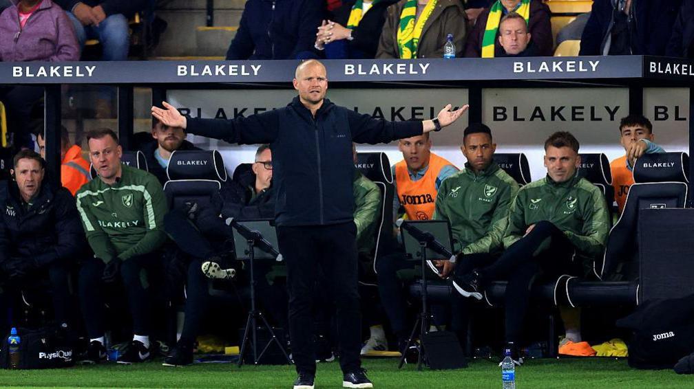 Liam Manning, in a dark blue track suit, stands in front of the dug out with his arms outstretched, seemingly expressing frustration at something happening on the pitch. There are players and coaches sitting in the dug out.