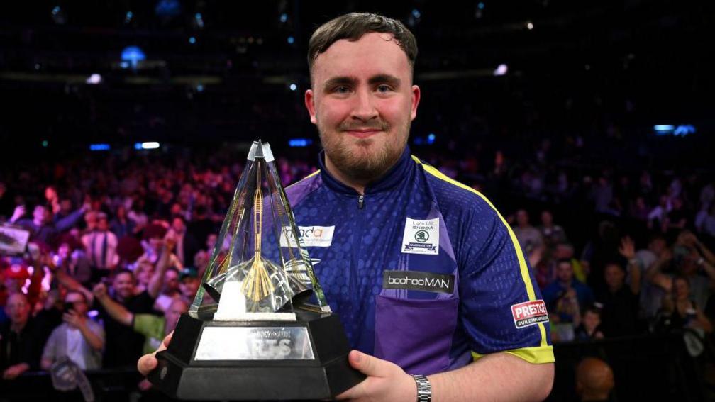 Luke Littler holds up the Premier League of Darts trophy.