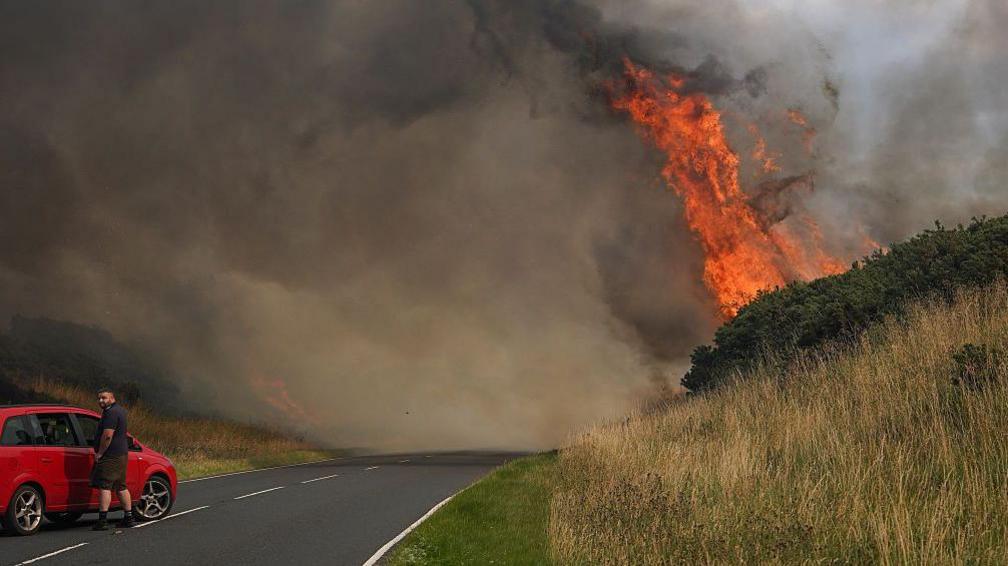 A man in a car watching flames burn nearby
