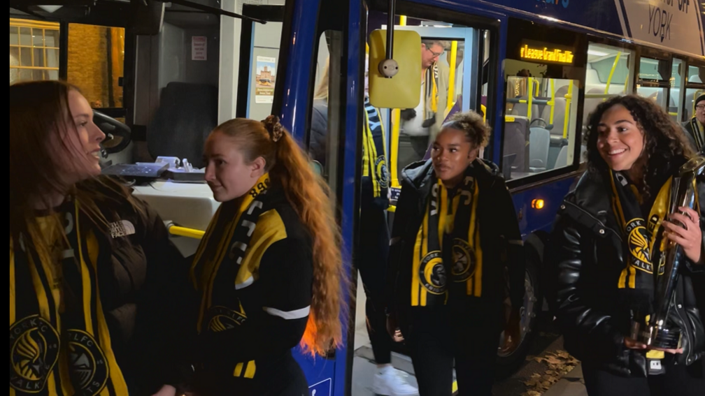 Four women wearing York Valkyrie scarfs and tops walk off a blue double decker bus.