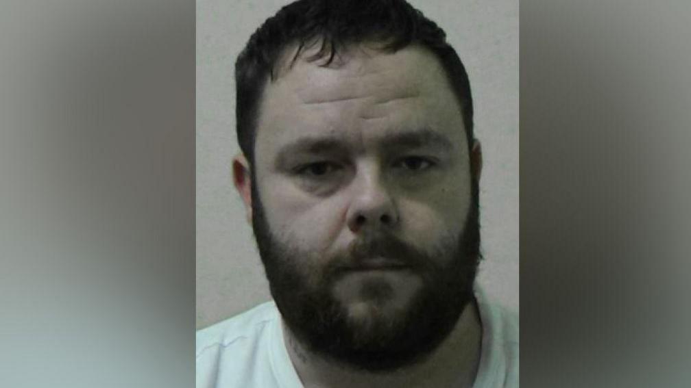 Mugshot of Binns. He has short dark hair and a thick dark beard and is wearing a white t-shirt.