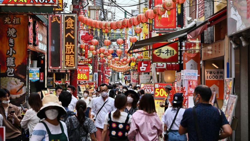 Crowds of people walk down a street in the Chinatown section of Yokohama. Many are wearing face masks and there are many, many shop signs and red lanterns hanging over the street.