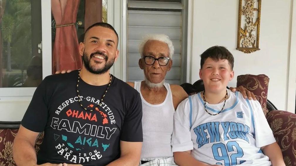 Three males sit alongside each other on a sofa in a house. From left to right they are: Jordan Murdoch, who appears aged in his 30s and is wearing a T-shirt with the words that read 'Ohana' and 'family'. In the middle is 98-year-old Hopeton Murdoch, Jodi's grandfather who is wearing glasses and a white vest. On the right is Jodi's son Julian who is wearing a New York branded basketball jersey.