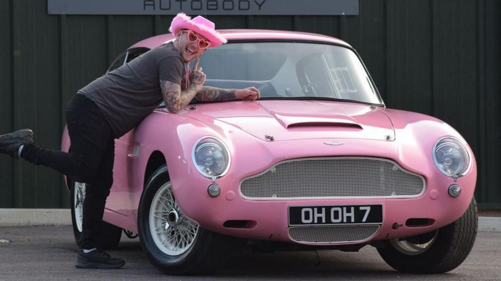 David Pearce-Martin, wearing pink Barbie hat and dark t-shirt and jogging pants draped over the bonnet of a pink 1967 Aston Martin DB6 he has restored. He's wearing pink heart glasses and is smiling to camera.