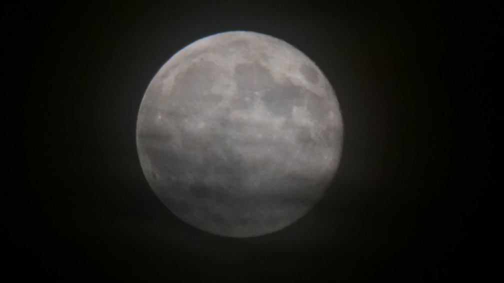 This image shows a full moon set against a dark night sky. The moon is captured in high detail, revealing surface features like craters and lunar maria (the darker plains). Its round shape and brightness stand out sharply against the surrounding darkness.