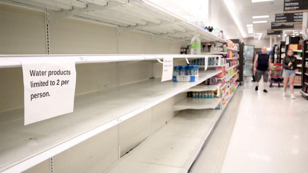 shelf in a supermarket, empty, with sign reading 'water products limited to one person'.