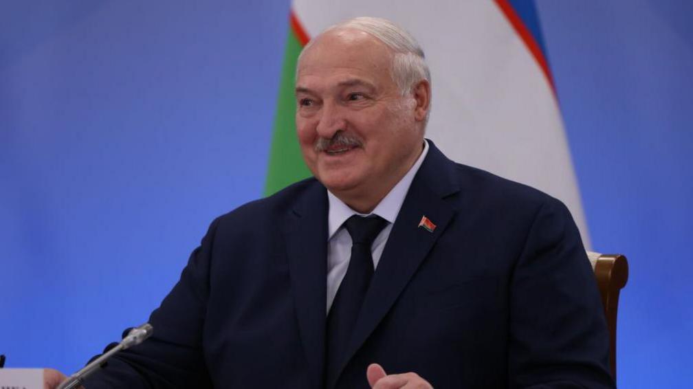 Alexander Lukashenko, sitting at a table with a mic in front of him. He is wearing a dark jacket and tie. A Belarusian flag is in the background. 