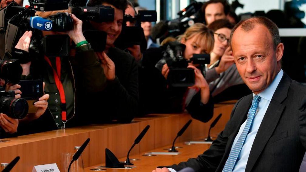 Merz sits in a suit and blue tie, staring at the camera, while TV cameras film him from behind