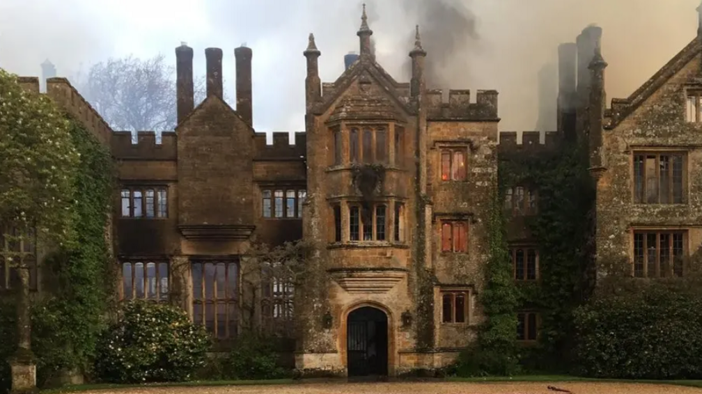 Flames can be seen in some of the windows of Parnham House, a stately home of up to three storeys, with tall chimneys, ornate pinnacles and crenellations. A fire service hose is laid out on the front lawn and drive and smoke is rising from the building.