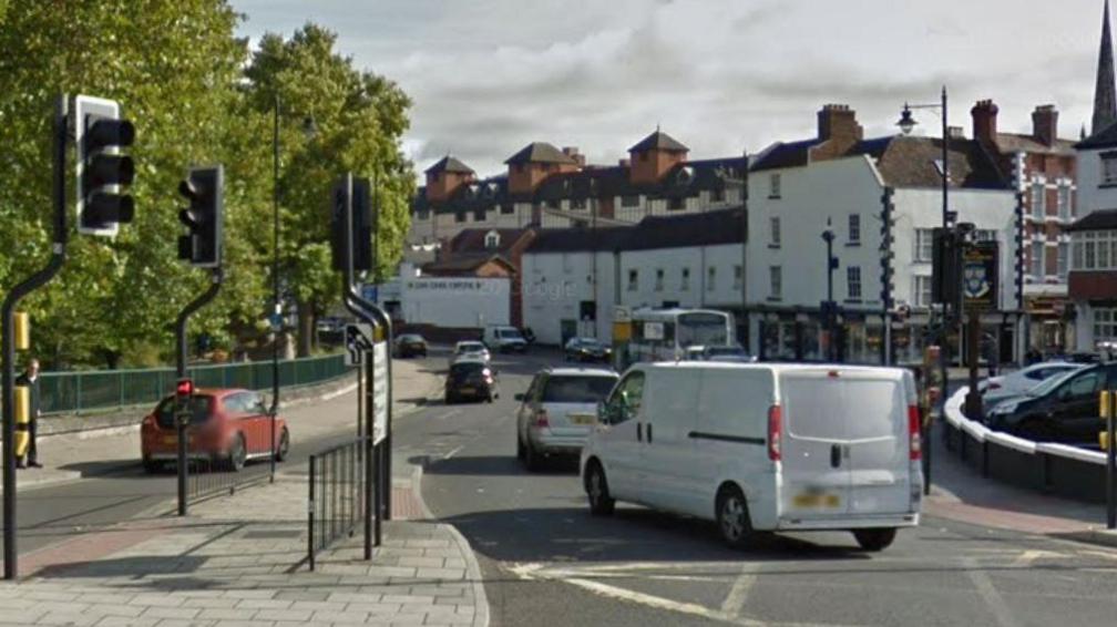 Cars and vans on a town centre street with traffic lights in the foreground and tall, white-walled buildings in the distance.