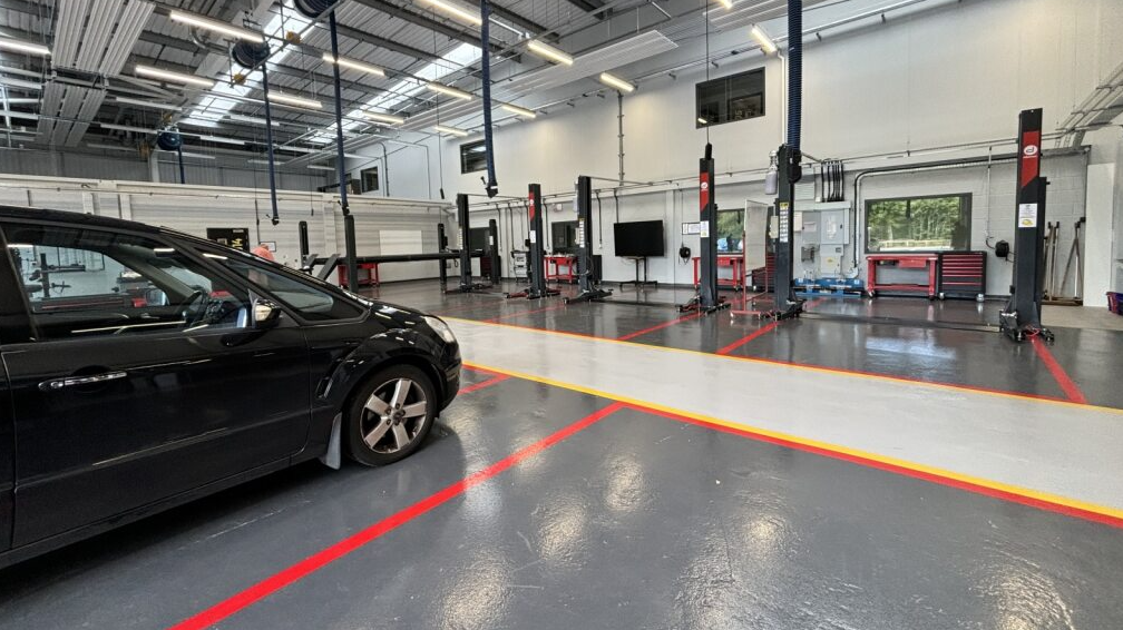 The image shows the insides of the working bays. The floor is painted grey with sections marked with red or yellow paints. A car is parked in a bay on the left with two or three empty bays in front of it. The building looks very new and well kept.