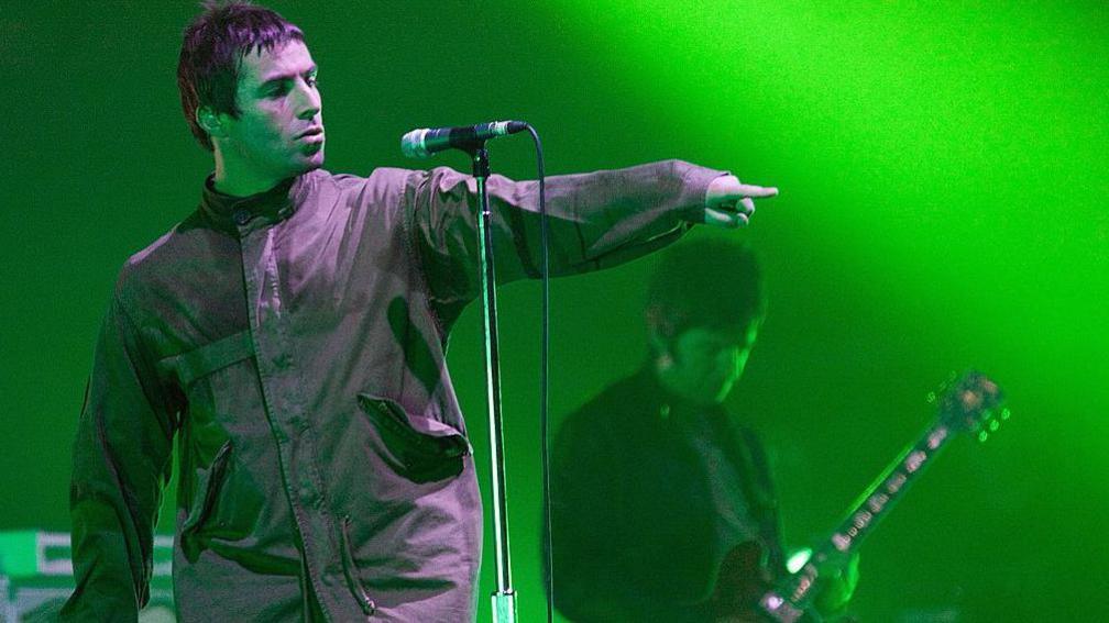Oasis lead singer Liam Gallagher in a parka jacket pointing at the crowd in front of a microphone with his brother and bandmate Noel behind him, face down playing a guitar on stage in 2009 with green lights lighting the stage