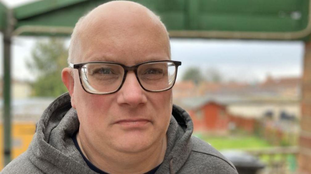 Kevin Slade wearing a grey hoodie and dark glasses. He is bald and looks middle aged. The background is a blurred garden. 