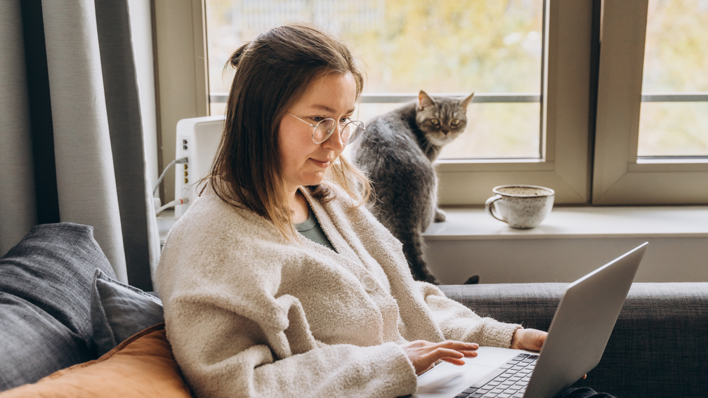 A young woman working from home on a laptop, sitting on a sofa and watched by a grey cat on the window sill. 