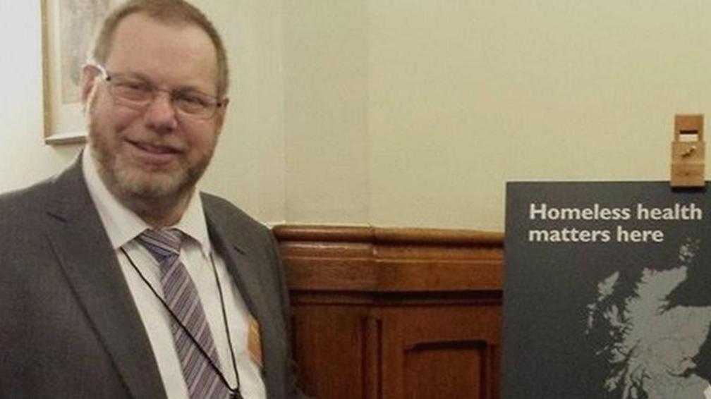 Nobby Clarke smiling at the camera in a grey suit jacket and white shirt and striped purple tie. On the right is a board that says 'Homeless health matters here' in white text.