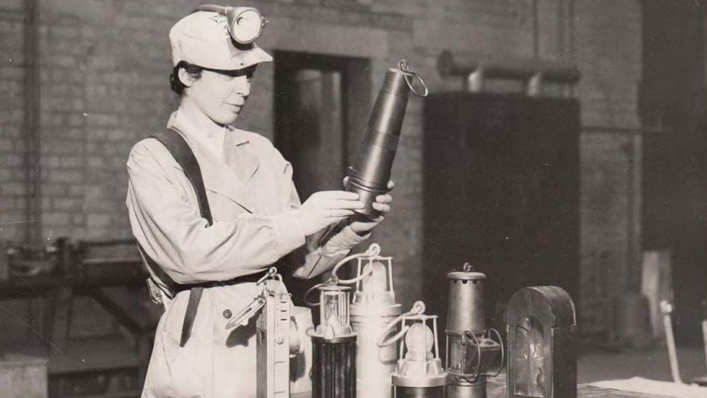 A woman stands in front of six miner lamps in what appears to be a work station. She holds one between her hands, appearing to inspect it. She is in a work uniform and wears a hat with a torch.