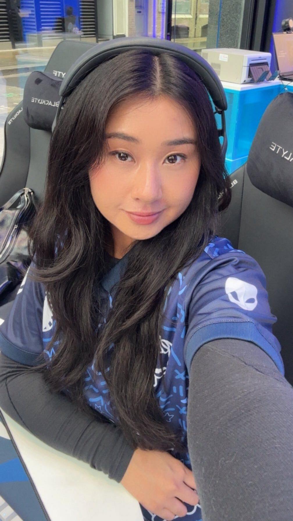 A woman wearing a gaming jersey and sitting in a black gaming chair. The person has long dark hair and is wearing a headset. In the background, there is a blue-lit gaming setup with a monitor, keyboard, and other equipment on a desk near a glass wall.