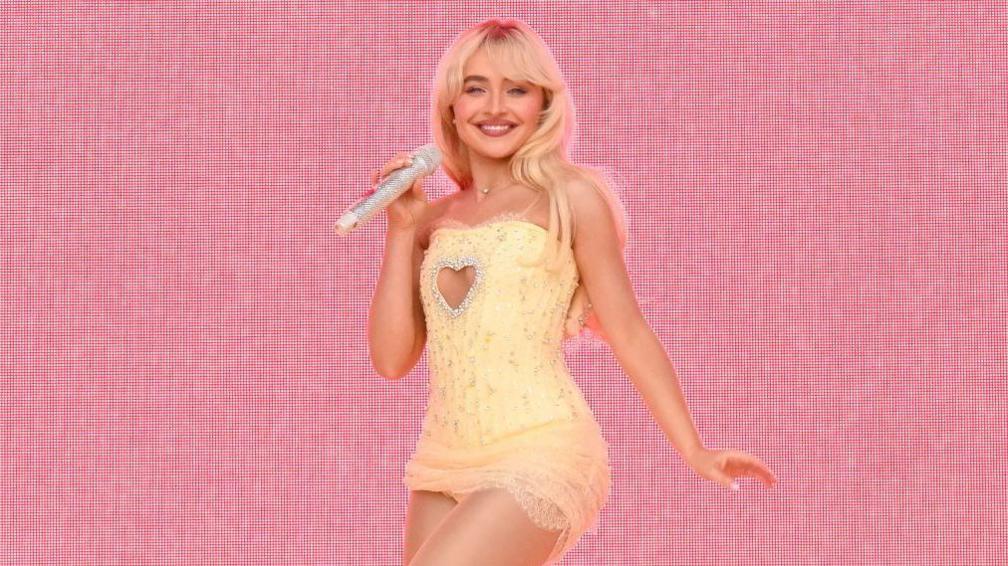 Sabrina Carpenter stands on stage during a performance. She is wearing a yellow dress and is holding a sparkly microphone.  