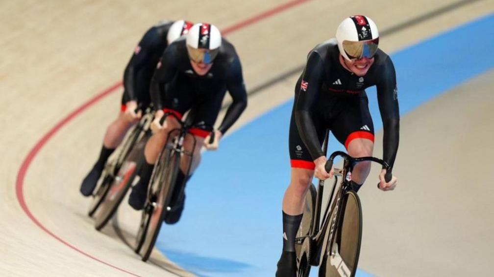 Action shot of Ed Lowe in his helmet and race outfit, gritting his teeth and looking up as he cycles the velodrome with teammates Hamish Turnbull and Jack Carlin behind him