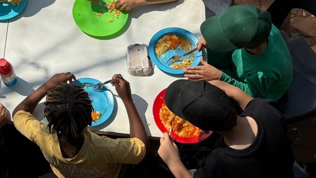 Children sit at three long tables which are placed in the schoolyard. They are eating lunch from brightly coloured plastic plates. The picture is taken from above, so you can only see the tops of their heads
