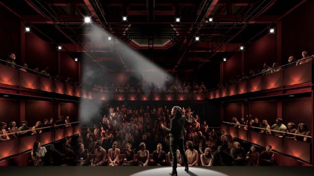 A computer-generated image of the auditorium of a theatre. A woman wearing all-black is standing on the stage under a spotlight, speaking to sell-out crowd.