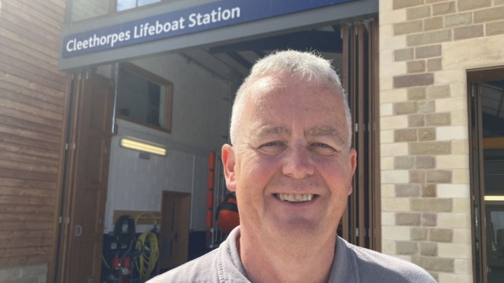 Man with grey hair smiling. He's wearing a grey t shirt and is standing in front of a stone building with wooden doors. There is a sign above the door which reads Cleethorpes Lifeboat station.