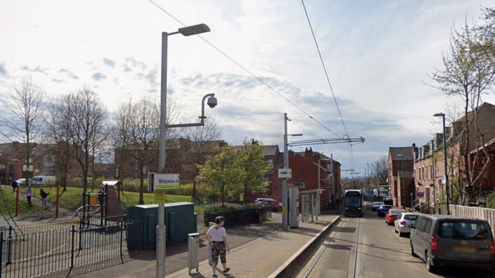 A Google Streetview image of Shipstone Street tram stop in New Basford in Nottinghamshire. The image shows a tram approaching the stop with cars parked on the right of the tram. There is a children's play area on the left of the the image, behind the tram stop.