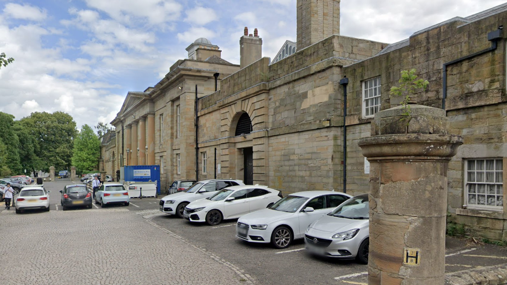 The exterior of Durham Crown Court. The building is made up of large sandstone bricks, and has a Georgian design with Roman columns at its entrance. There are several cars parked outside on a cobbled area of road. 