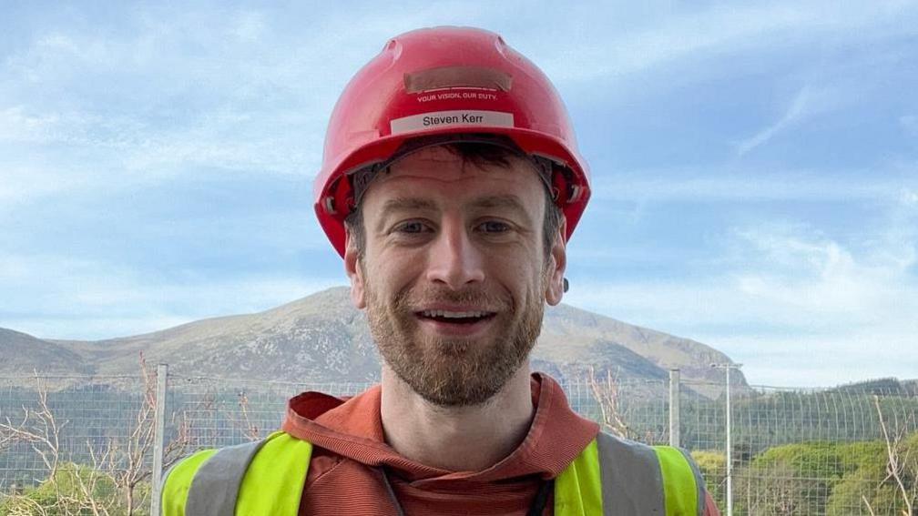 Mr Kerr has dark brown hair and facial hair he is smiling at the camera as he wears a red hard hat, a red hoodie and a yellow hi-vis. Behind him is a mountain range and a metal fence.