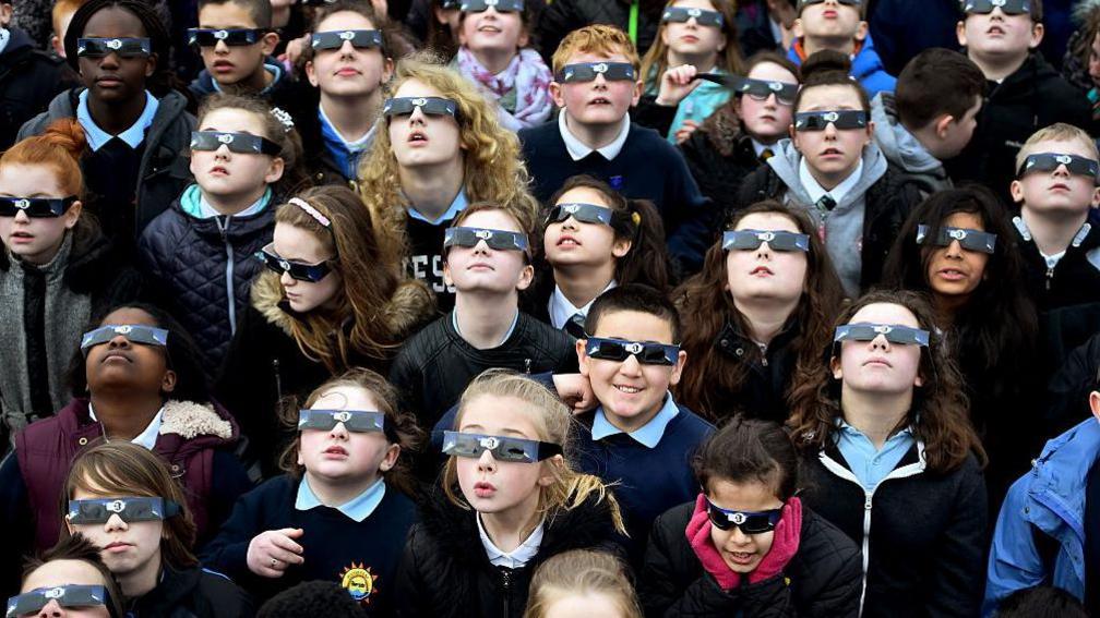 School pupils all wearing eclipse glasses and looking up towards the sky. The group includes a mix of boys and girls and they are wearing a navy blue school uniform.
