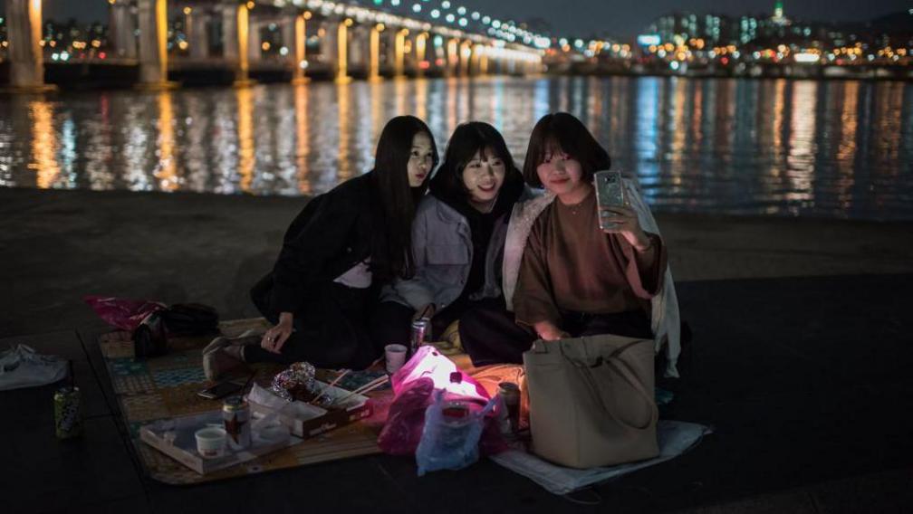 A group of women smile and look at a mobile phone one of them is holding up as they pose for a selfie before the Banpo bridge on October 13, 2016. They are sitting on a blanket on a patch of grass, dressed in warm clothes. In front of them are food containers and their bags. Behind them is a lake and the Seoul city skyline.
