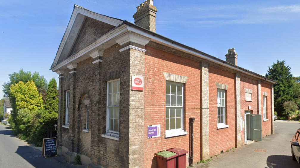 A general view of the exterior of a post office building. The single-storey building has a mixture of red and yellow brick. It has several large windows and a post office sign can be seen on its side.