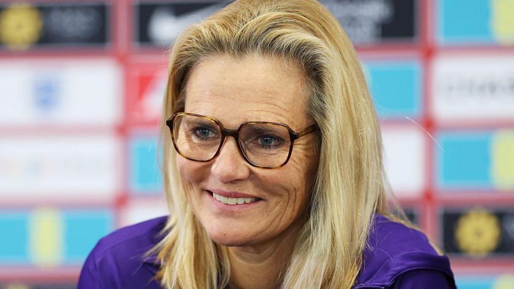 England boss Sarina Wiegman smiles at a press conference. She is wearing glasses and a purple England top. 