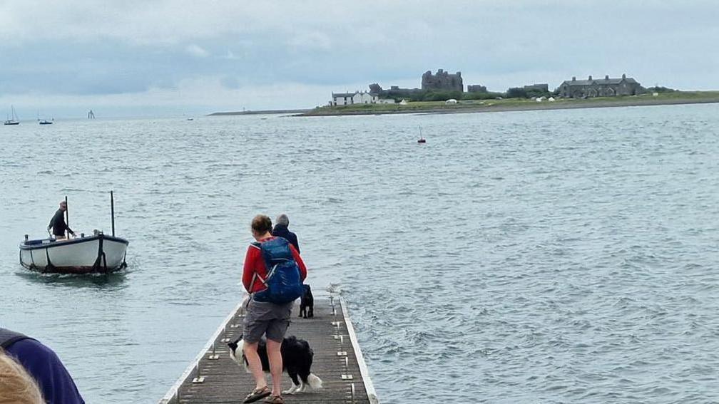 A pier stretching out to the sea with people and a border collie waiting for a boat. The boat is a large rowing boat to the left and a man is steering it. In the background, Piel Island can be seen - it has rocky ruins and a few small buildings.