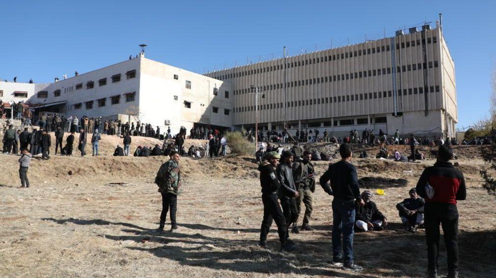 People gathering outside Saydnaya Prison, which is a big white building.