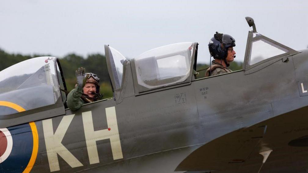 Doreen Allen, in the passenger seat of a grey Spitfire, is wearing an olive green flight suit and waves at the camera