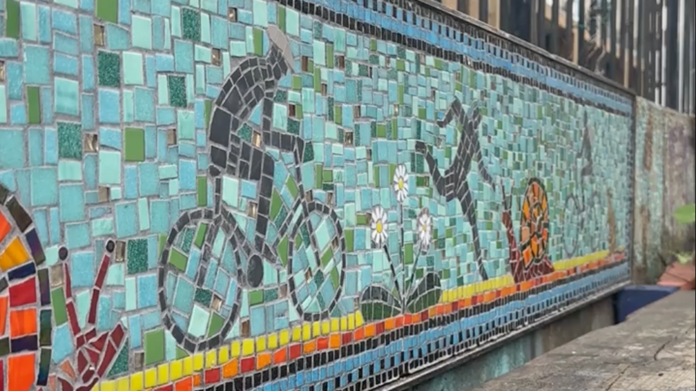 A mosaic is featured behind a wooden bench. The colours are blue, yellow, orange and black and feature figures running and cycling along the canal, next to plants and snails