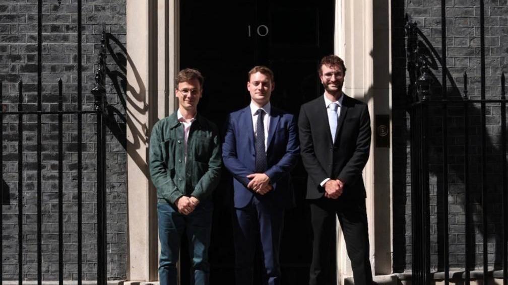 Three men dressed smartly with hands crossed in front of them standing in front of the Number 10 building in London.