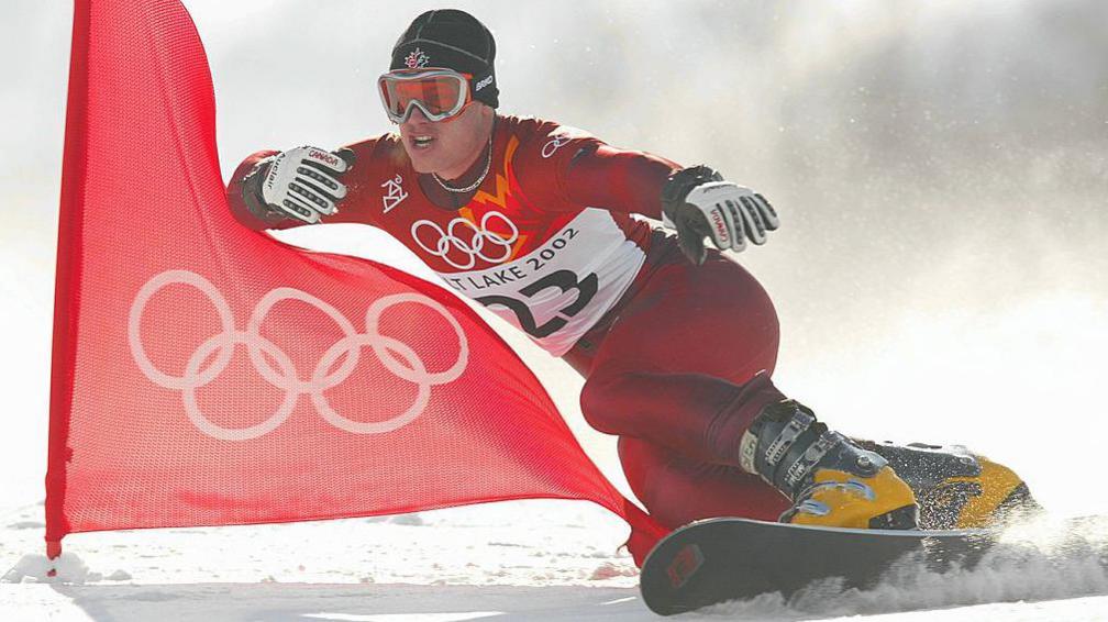 Ryan Wedding snowboarding in 2002 at the Winter Olympics in Salt Lake, Utah, USA. He wears a red, Lycra sporting top and trousers featuring the Olympic rings and the Canadian maple leaf, along with red goggles, a black hat, thick gloves and yellow boots. He is snowboarding passed a red flag with the five Olympic rings.