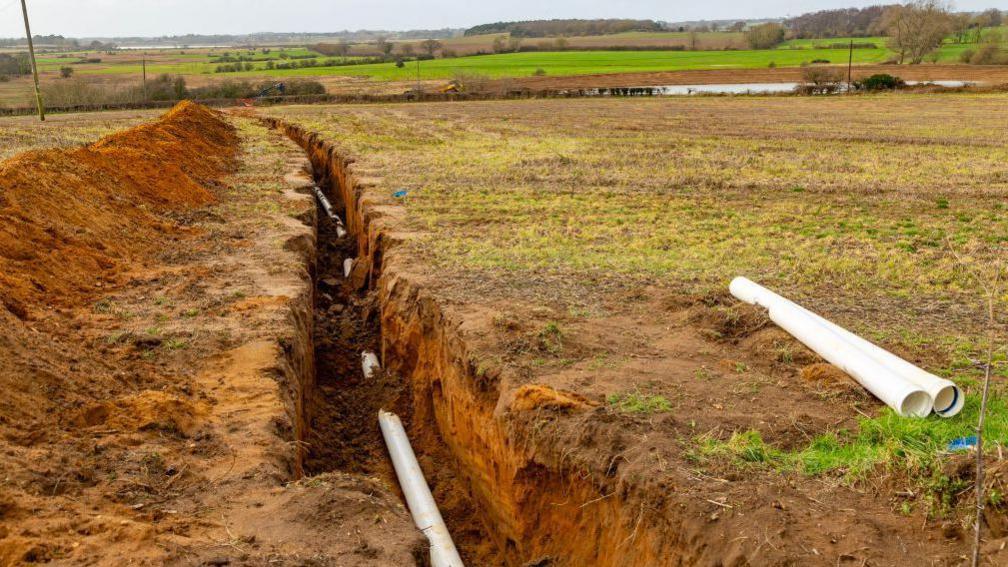 Farm irrigation water supply pipeline excavated in sandy soil, Shottisham, Suffolk, England, UK reservoir in distance.