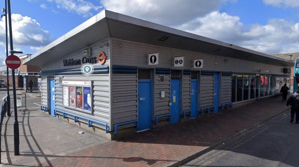 Waltham Cross bus station, a grey building with blue doors, glass windows and blue metal railings around the bottom. There is a no entry sign to the left and a bus to the right. There are signs to three toilets. 