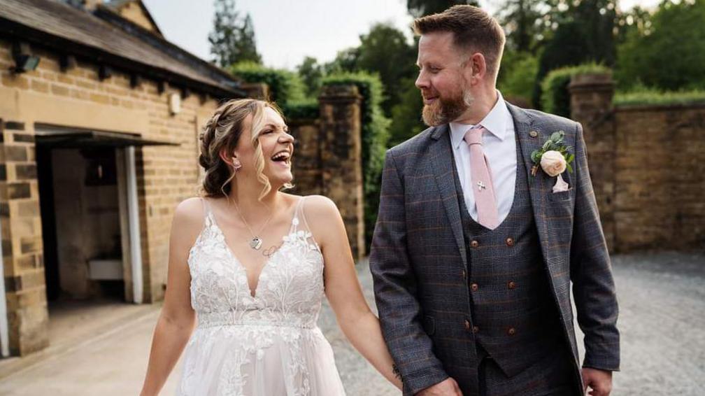 Charlotte and Dave on their wedding day. They are holding hands and are looking at each other and smiling. Charlotte is in a white lacey dress and Dave is in a grey checked suit with a pink tie. They are outside a wedding venue.
