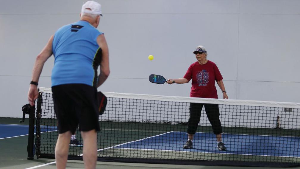 Two older people in leisure wear playing pickleball on an outdoor court. There is a net and the court floor has markings. 