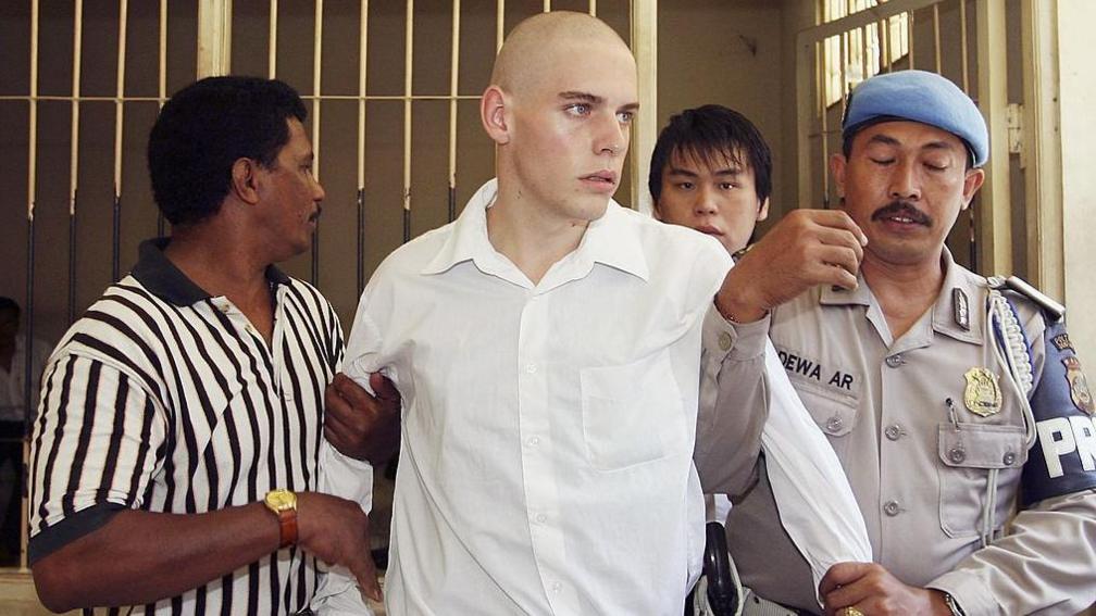 Indonesian police escort Matthew Norman and Si Yi Chen, who are both young men, to Denpasar District Courtroom for their sentencing trial. Norman wears a white shirt in the foreground. His arms are being held by Indonesian prison guards. A barred cell is open in the background.