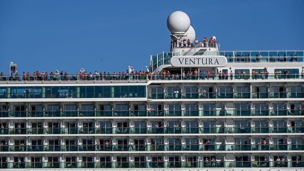 Passengers on Ventura, some are on the balconies of their cabin and some at the rooftop of the ship on a clear day. The VENTURA sign is seen. 